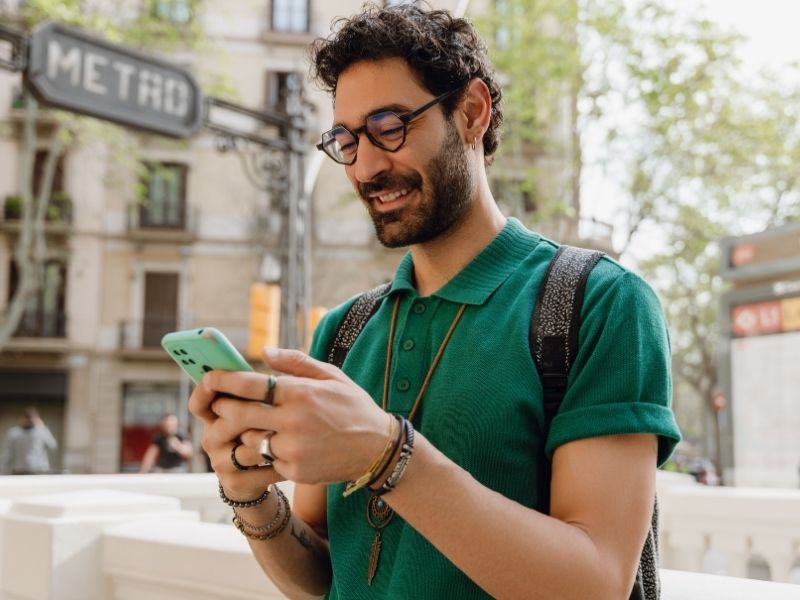 Hombre sonriendo con su teléfono en una calle, junto a un letrero de metro